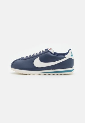 Marineblauwe Nike lage sneaker met witte swoosh, witte veters en geribbelde witte zool met blauwe streep, zijaanzicht op witte achtergrond.