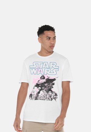 Young man wearing a white Star Wars T-shirt with characters and logo looking to the side against a plain gray background.