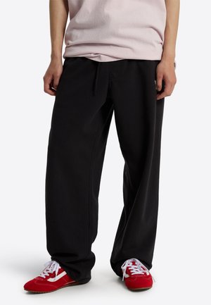 Person iført løstsiddende sorte bukser, lyserød skjorte og røde sneakers med hvide snørebånd, stående mod en ensfarvet baggrund.