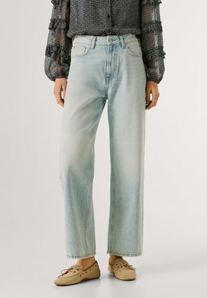 Persona vistiendo jeans de pierna ancha de color azul claro, una blusa de manga larga oscura con estampado y mocasines beige con cordones, de pie sobre un fondo claro.