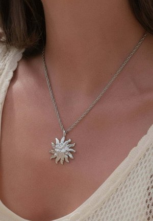 Silberner sonnenförmiger Anhänger an einer verdrehten Kettenhalskette, getragen auf der Brust einer Frau, mit einem cremefarbenen, strukturierten Oberteil sichtbar an der Schulter.