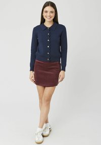 Cardigan bleu marine à ourlet festonné avec des boutons dorés, associé à une mini-jupe en velours côtelé bordeaux et des baskets blanches. Texture de tissu lisse.