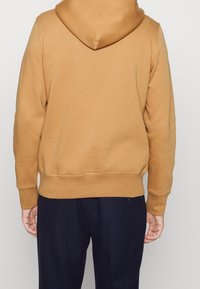 Sudadera marrón hecha de una tela suave. Presenta una capucha, puños y dobladillo acanalados, y un corte holgado. Combinada con pantalones oscuros.