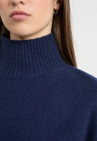 Granatowy, dzianinowy sweter z golfem o prążkowanej strukturze; noszony blisko szyi, z dopasowanym krojem i widocznymi detalami przeszycia.