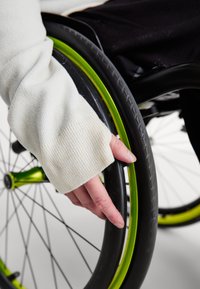 Λευκό, υφασμάτινο μανίκι ρούχου πάνω σε ένα χέρι που κρατάει ένα μαύρο ελαστικό αναπηρικής καρέκλας με πράσινη στεφάνη, αναδεικνύοντας τον στρογγυλό του σχήμα και τις ακτίνες.