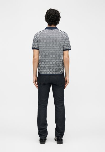 Homme aux cheveux bouclés portant un polo à manches courtes à motifs, un jean foncé et des chaussures noires, debout de dos devant un fond uni.