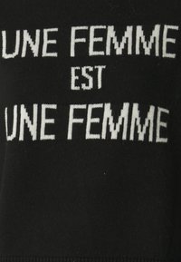 Svart stickad plagg med vitt text som säger "UNE FEMME EST UNE FEMME", med en framträdande design i fet stil.