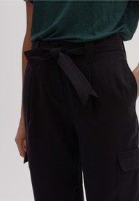 Pantalon noir en tissu texturé, avec une ceinture nouée à la taille. Comprend des poches latérales et une coupe ample.