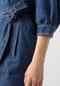 Denimkjole med belte i midjen, puffermer og knappelukking, i en mellomblå nyanse med kontraststing.
