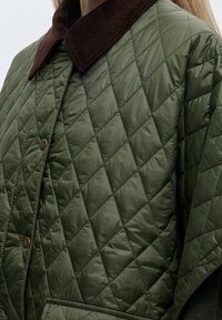 Femme portant une veste matelassée vert olive avec un col en velours côtelé marron et des boutons-pression métalliques.