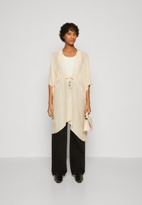 Lett, beige kimono-stil cardigan med åpen front, sidespalter og knytedetalj, kombinert med en enkel hvit topp og svarte vide bukser.