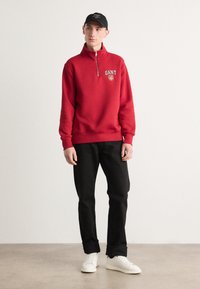 Sweatshirt roșu cu guler înalt și fermoar pe jumătate, cu un logo. Asortat cu pantaloni negri și adidași albi, pe un fundal neutru deschis.