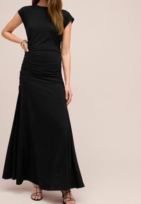 Femme portant une longue robe noire froncée avec des manches courtes et des sandales noires à lanières, accessoirisée de bracelets en or.