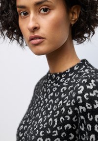 Giovane donna con capelli corti e ricci, che indossa un maglione a pois leopardati in bianco e nero e un piccolo orecchino ad anello d'oro.