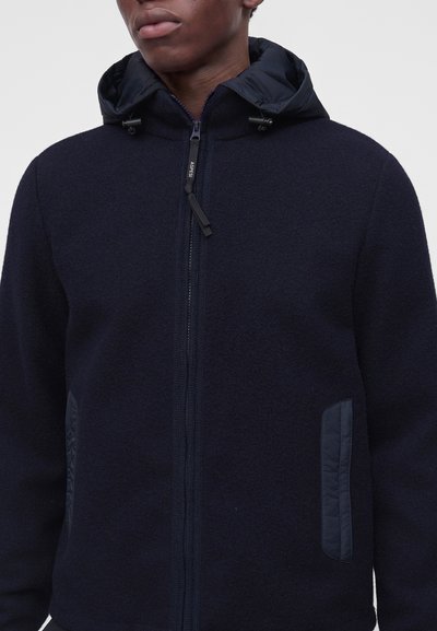Homme portant un sweat à capuche zippé bleu marine foncé avec des accents matelassés sur les poches et une capuche avec cordon, sur un fond clair uni.