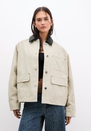 Beige Canvas-Jacke mit schwarzem Kragen, Frontdruckknöpfen, zwei aufgesetzten Taschen und lockerem Schnitt. Kombiniert mit blauen Leoprint-Jeans.