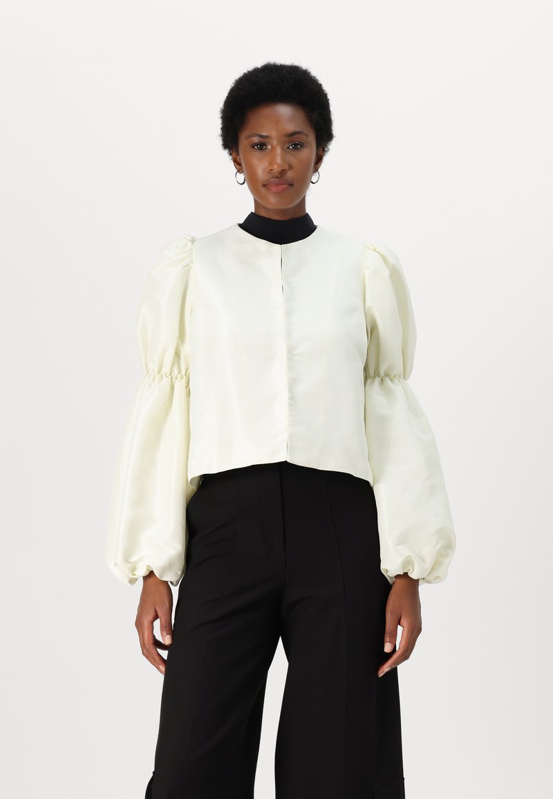 Blouse en satin crème coupée, avec des manches bouffantes et un col montant noir. Présente une fermeture à boutons à l'avant et une texture lisse. Associée à un pantalon noir.