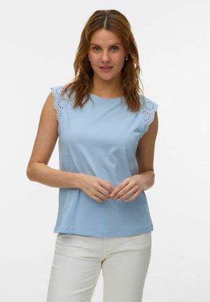 Kvinde med bølget brunt hår iført en lyseblå ærmeløs top med blonderkanter og hvide bukser, stående mod en ensartet hvid baggrund.