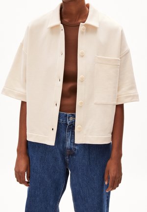 Personne portant une chemise crème à manches courtes boutonnée par-dessus un haut marron et un jean bleu taille haute, debout avec les bras détendus.