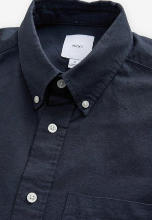 Jūras zilā blūze ar strukturētu apkakli, baltām pogām un mazu krūšu kabatu. Gluda auduma ar smalku tekstūru. Redzama zīmola etiķete.