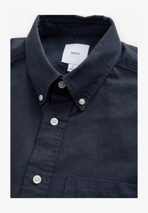 Jūras zilā blūze ar strukturētu apkakli, baltām pogām un mazu krūšu kabatu. Gluda auduma ar smalku tekstūru. Redzama zīmola etiķete.