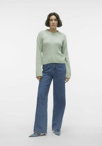 Pulover ribbed de culoare verde deschis, cu mâneci lungi, asortiat cu blugi albastri cu talie înaltă și picioare largi, purtând pantofi cu toc albastri, ascuțiți.