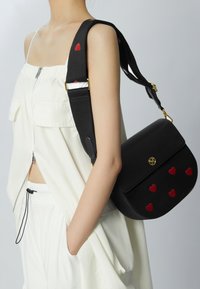 Černá kožená kabelka se zlatými detaily a červenými aplikacemi ve tvaru srdce. Má černý popruh s červenými srdci a zipové zavírání.