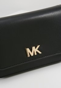 Czarny skórzany portfel o gładkiej teksturze, ze złotym logo "MK". Zawiera wyraźne przeszycia oraz elegancki, kompaktowy design.