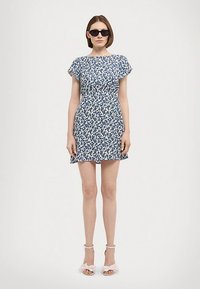 Φλοράλ φόρεμα με μπλε και λευκό μοτίβο, κοντά μανίκια, εφαρμοστή μέση, φουσκωτή φούστα, φορεμένο με μαύρα γυαλιά ηλίου και ανοιχτόχρωμα παπούτσια.