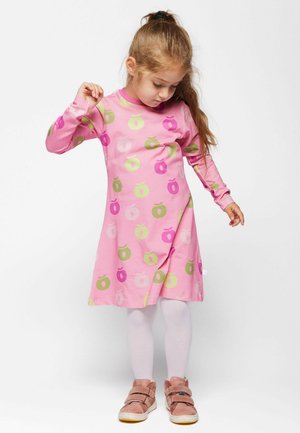 Mlada djevojka s dugom kosom u ružičastoj haljini s uzorkom jabuka, bijelim tajicama i ružičastim cipelama, gleda prema dolje i podiže jednu ruku.