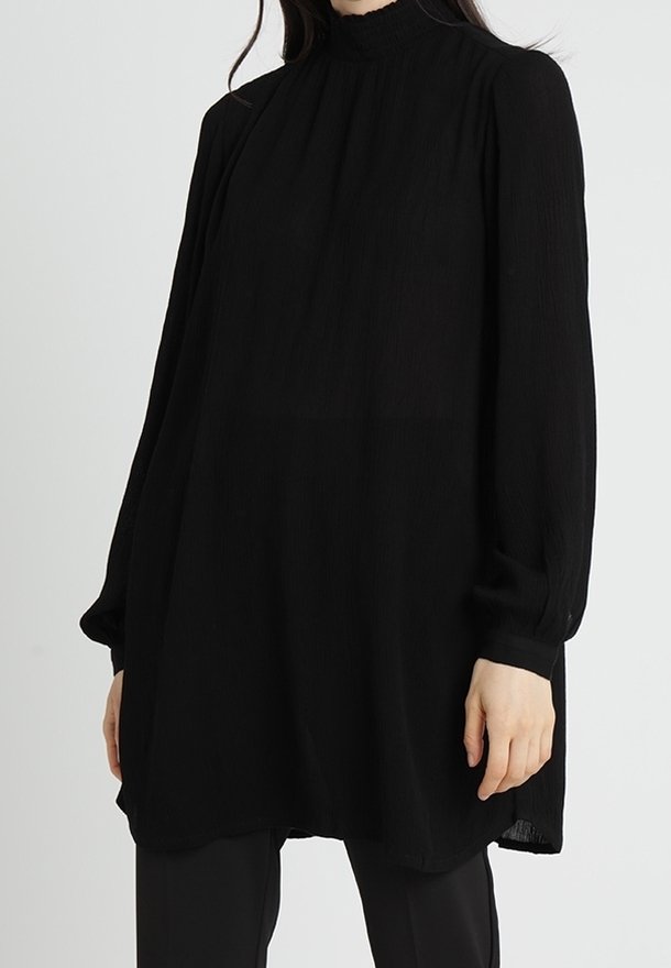 Femme portant une tunique noire longue et ample à col montant et manches longues, associée à un pantalon noir sur un fond clair uni.