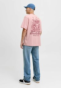 Lyserød oversized t-shirt med et detaljeret grafik på ryggen, parret med lyseblå jeans og sorte sneakers med hvide detaljer.