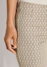 Linkerhand ligt rustend naast een beige en grijs houndstooth-patroonrok of -broek tegen een neutrale achtergrond.
