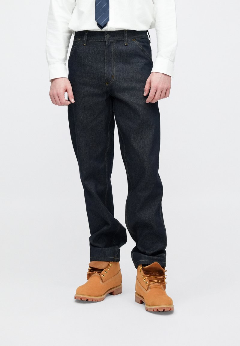 Dunkelblaue Jeans aus Denim mit oranger Naht, Knopfverschluss und geradem Bein. Kombiniert mit hellbraunen Schnürstiefeln.