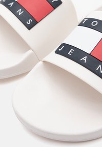 Valged libisemise sussid, millel on tekstureeritud ülemine osa ja julge logo punases, valges ja mustas. Pehme tallapadi suurendab mugavust.