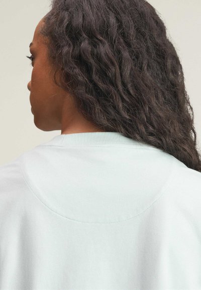 Frau mit langen, lockigen, dunklen Haaren, die ein hellblaues Rundhalsshirt trägt, von hinten und im Seitenprofil vor einfarbigem Hintergrund dargestellt.