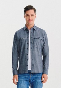 Mężczyzna stoi, ubrany w szary, zapinany na guziki koszulę na białej koszulce oraz niebieskie dżinsy, na jednolitym białym tle.