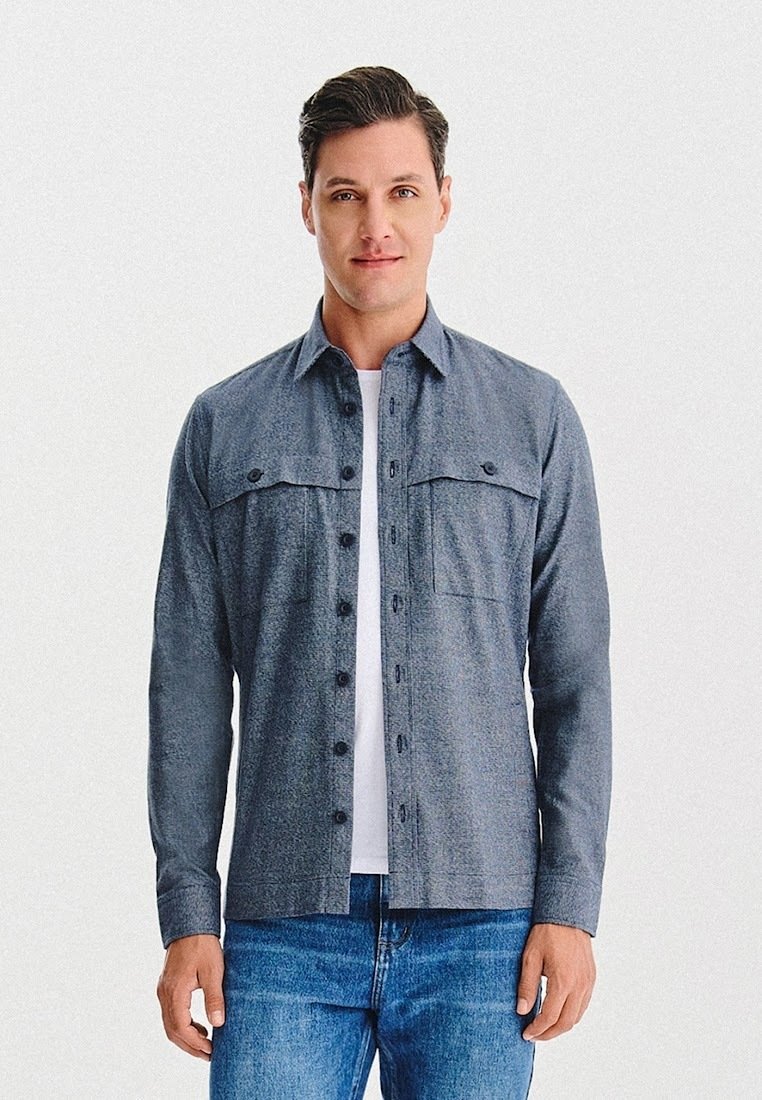 Mężczyzna stoi, ubrany w szary, zapinany na guziki koszulę na białej koszulce oraz niebieskie dżinsy, na jednolitym białym tle.