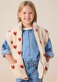Gilet en polaire de couleur crème avec des motifs de cœurs rouges, associé à une chemise en denim bleu clair et à un jean présentant des accents de cœur similaires.