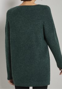 Pull en maille oversize de couleur vert foncé, avec une texture côtelée, un col large et des manches longues, couvrant les hanches.