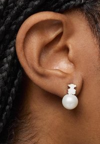 Pendientes de estudiod en forma de oso blanco con un colgante de perla redonda, sobre un peinado trenzado oscuro y tonos de piel naturales.