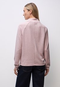 Pull rose clair, texturé, à col montant, avec des manches raglan et un ourlet ajusté. Associé à un jean en denim foncé, montré de dos.