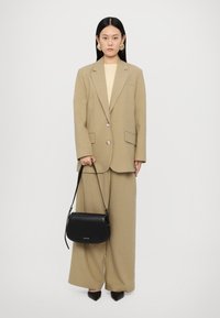 Blazer beige su misura con bottoni, pantaloni a gamba larga coordinati, top chiaro e borsa a tracolla nera. Modello in tacchi a punta neri.