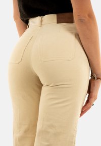 Pantaloni in corda color crema con vita alta, texture a righe verticali, tasche posteriori e un'etichetta del marchio in pelle marrone.