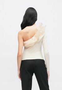 Asimetriškas smėlio spalvos viršus su viena ilga rankove ir raukiniais permatomame peties dizaine, derinamas su juodomis kelnėmis. Lygus paviršius.