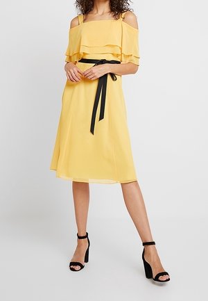 Vrouw draagt een gele off-shoulder midi-jurk met ruches en een zwarte tailleband, gecombineerd met zwarte sandalen met hoge hak en enkelbandje.