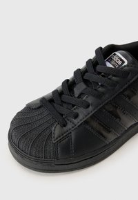 Μαύρο αθλητικό παπούτσι με υφή δέρματος, καουτσούκ σόλα και σχέδιο με τρεις ρίγες. Διαθέτει στρογγυλή μύτη και κλείσιμο με κορδόνια.