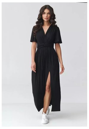 Kobieta o długich falowanych włosach, ubrana w czarną, maxi sukienkę z dekoltem w serek, krótkimi rękawami i rozcięciem z przodu, w białych tenisówkach, stojąca na białej podłodze.