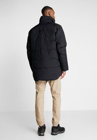 Black puffer jacket with a high collar, quilted design, and a slightly extended back. Paired with light beige pants and black shoes.