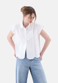 Weißes kurzärmliges Blusenshirt mit Kragen, gewelltem Saum und glatter Textur, kombiniert mit hellblauen, lockeren Jeans.